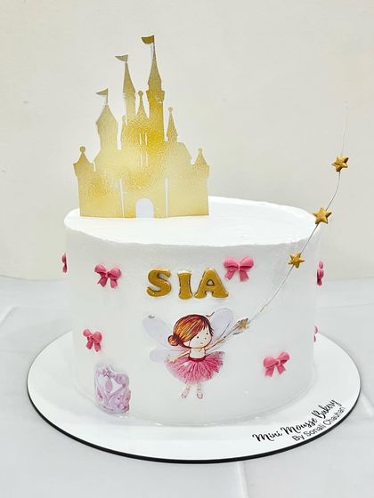 A fairytale cake for a little princess. This design features a fairy illustration, a gold castle topper, and tiny pink bows for a touch of magic.