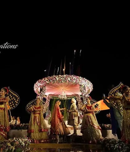 A wide-angle shot of a beach wedding Jaimala, capturing the entire magnificent setup. The stage, performers, and fireworks come together to create a scene of epic proportions.