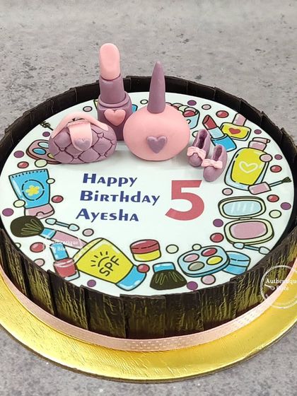 A cake for a little makeup freak. The top is decorated with edible illustrations of makeup products, with 3D fondant lipstick, nail polish, and a purse.