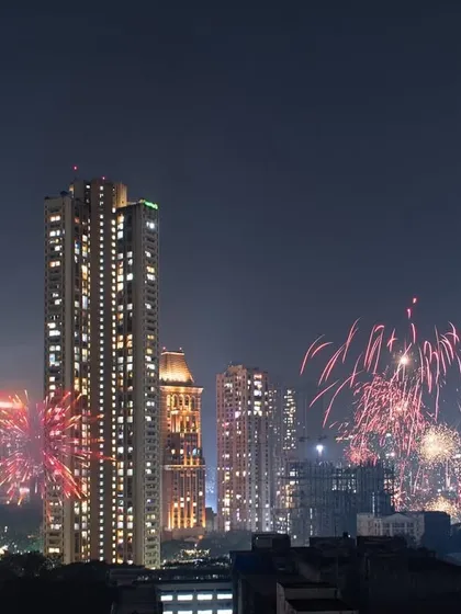 Red and pink Diwali fireworks burst across the night sky, illuminating the dense urban landscape of Mumbai's residential buildings.