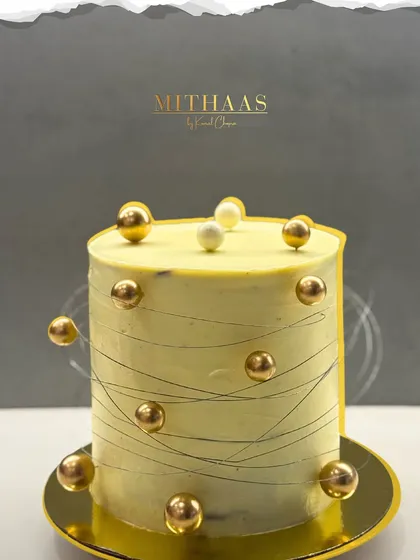 A tall, minimalist cake with a pale yellow frosting, decorated with a cascade of gleaming gold spheres. The fine, thread-like lines etched into the side add a subtle, modern texture to this sophisticated design.