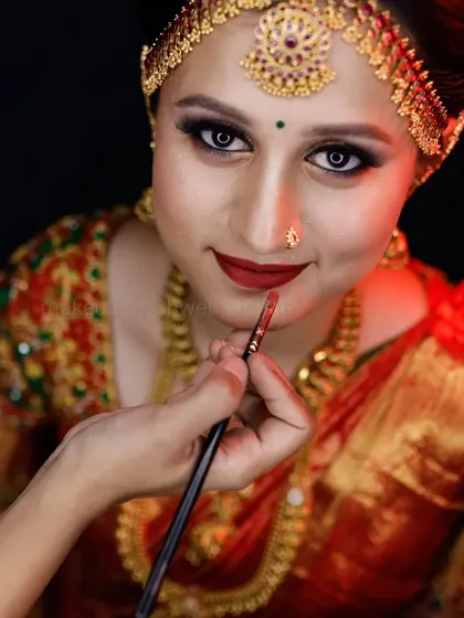A classic muhurtham look featuring a bright red lip and defined eyes. The traditional matha patti and heavy gold jewellery are perfectly balanced with the makeup, and the bridal braid with fresh flowers is a timeless choice.