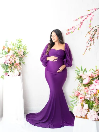 A vision in violet. The mother-to-be looks serene in a beautiful purple gown with puff sleeves, standing in a studio set decorated with lovely floral arrangements.
