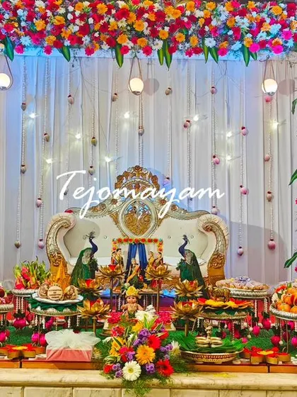 A vibrant baby shower setup with a colorful floral arch and a central seating area. The plate decoration is flanked by two traditionally dressed dolls.
