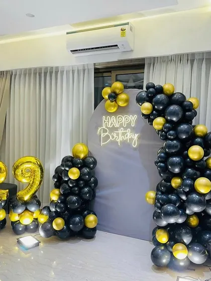 A sleek and modern 29th birthday decoration for a home setting. This design includes an elegant balloon arch in black and gold against a simple grey backdrop, with matching foil number balloons.