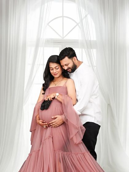 A tender moment where the couple holds a tiny pair of black baby shoes against the bump. This studio shot uses a soft, bright background to create a gentle and loving mood.