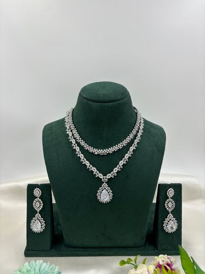 A graceful double-layered AD necklace. The top layer is a simple floral chain, while the bottom layer features a pear-shaped pendant. It is available in white, pink, green, and blue stones.