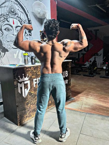 A back double bicep pose in front of the gym's reception. The Hanuman mural in the background adds to the powerful vibe.