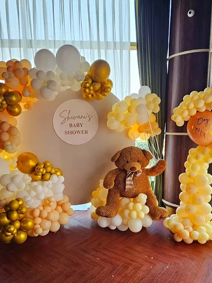 A teddy bear themed baby shower for Shivani. The decor features a warm color palette of mustard yellow, gold, and white balloons, with a large teddy bear as the centerpiece.
