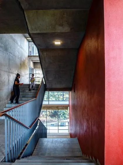 A view of the primary circulation spine at the JSW School of Public Policy, showcasing the use of raw concrete, a bold red feature wall, and a generous staircase to create a dynamic and engaging institutional interior.
