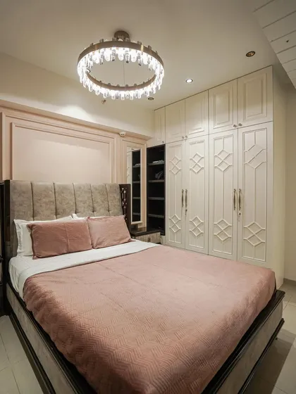 A luxurious bedroom with blush pink walls, ornate white wardrobes, and a crystal ring chandelier.
