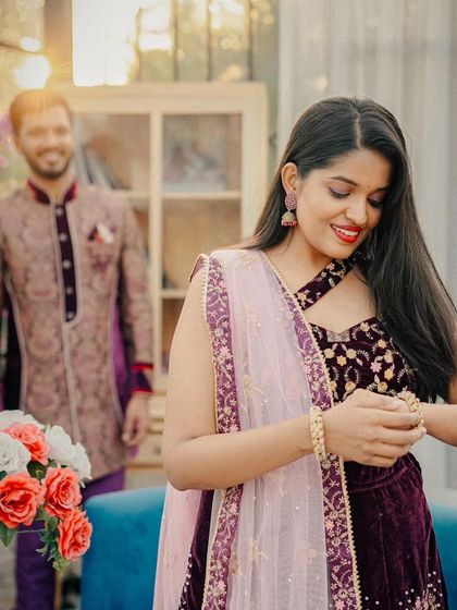 A candid moment during a pre-wedding shoot. The focus is on the bride-to-be in her gorgeous velvet lehenga, with her partner in the background.