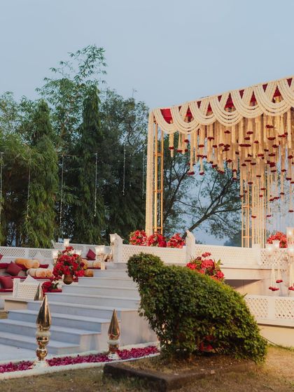 A wide view of the outdoor wedding setup, showing the tiered mandap structure against a lush green backdrop. The seating arrangement on the steps provides guests with an excellent view of the ceremony.