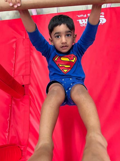 A young boy in a Superman shirt hangs from the bar, focused and determined. This exercise is excellent for developing the grip and upper body strength that is fundamental to gymnastics.