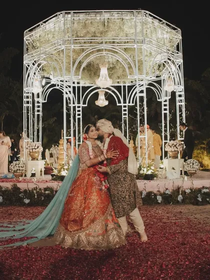 The couple shares a kiss in front of their beautiful wedding mandap, a perfect moment framed by the elegant structure and a carpet of scattered rose petals.