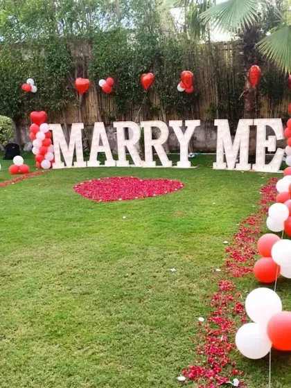 Agar aap kisi ko propose karna chahte hain, toh hum uske liye bhi special outdoor setup taiyar karte hain. Yeh 'MARRY ME' light letters, red aur white balloons, aur gulab ki pankhudiyon se sajaya gaya ek perfect proposal decoration hai.