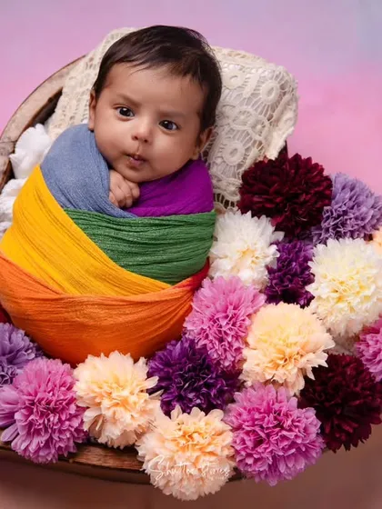 This awake newborn is wrapped in a rainbow of colors, looking curiously at the camera from a bowl of colorful pom-poms.