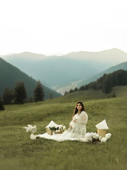 A breathtaking portrait in the mountains. The mother-to-be sits on a picnic blanket in a vast green meadow, with layers of mountains stretching out behind her, creating a sense of peace and grandeur.