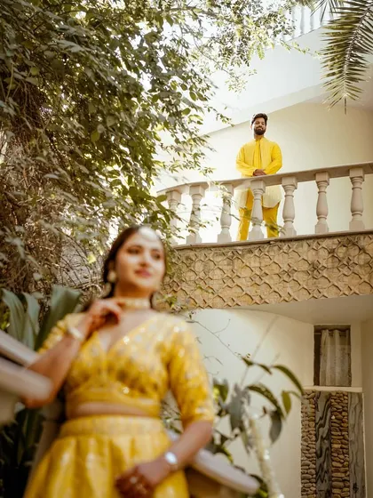 A creatively framed shot with the groom on an upper balcony looking down towards his bride. This composition adds depth and a storytelling element to their Haldi ceremony photos.
