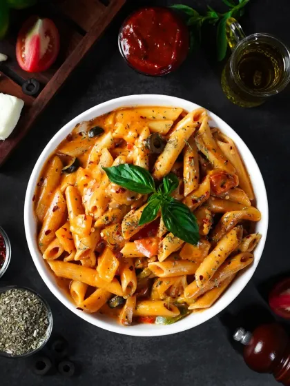 A bowl of creamy penne pasta in a rosé sauce, shot from above in a dark and moody style. The composition with fresh ingredients surrounding the bowl highlights the dish's flavors.