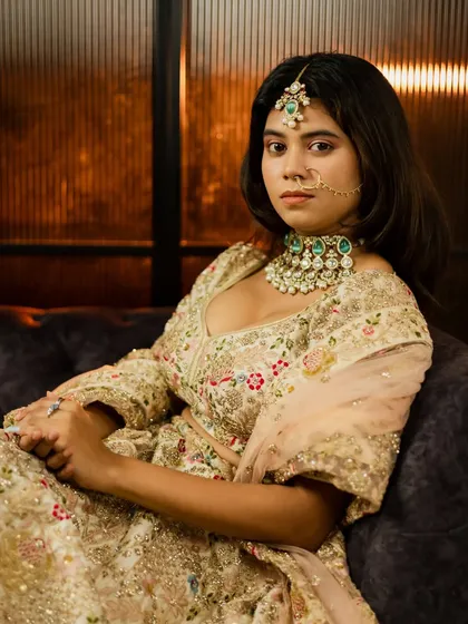 A full-length bridal portrait in an elegant indoor setting. The bride is seated, showcasing her magnificent embroidered lehenga and stunning Polki jewelry.