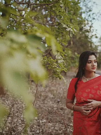 A woman in a red saree walking down a rustic path, captured from a distance with foliage creating a natural frame.