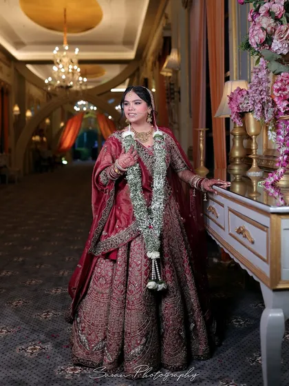 An elegant portrait of the bride in a grand hallway, her maroon lehenga and garland creating a regal look.
