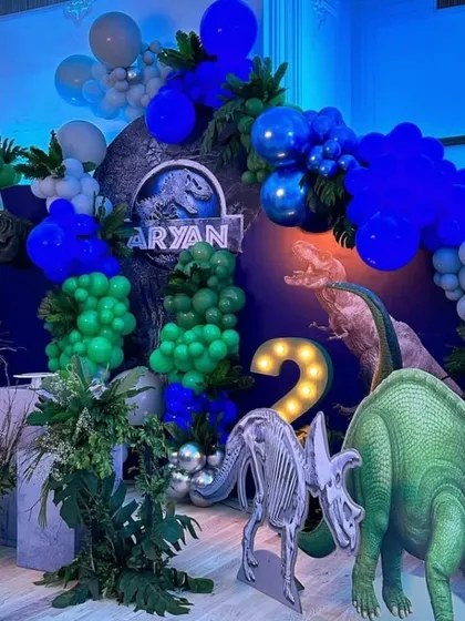 A roaring good time with this dinosaur-themed birthday party. The backdrop features a Jurassic Park-style logo, lush greenery, and large dinosaur props.