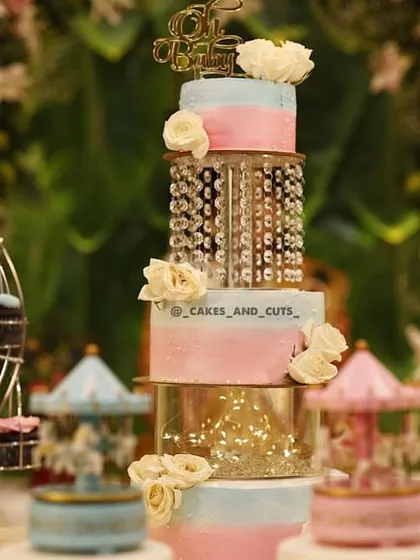 A close-up of a multi-tier baby shower cake with pink and blue ombre frosting, fresh white roses, and clear tiers filled with crystals and fairy lights.