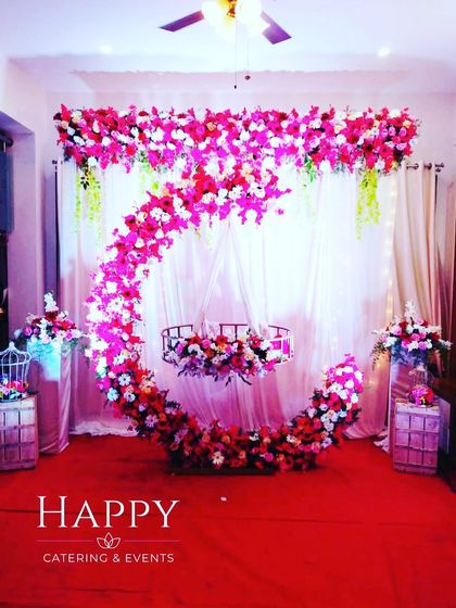 A vibrant naming ceremony stage featuring a crescent moon cradle decorated with bright pink and red flowers. The white curtain backdrop makes the floral colors stand out beautifully.