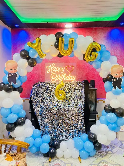 A Boss Baby themed birthday setup for Yug's 6th birthday. The decor includes a blue, black, and white balloon arch, a silver shimmer backdrop, and foil name balloons.