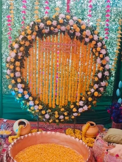 A beautiful Haldi setup with a large brass urli filled with marigold petals. The circular backdrop is made of marigold strings and framed with yellow and white flowers, set against green drapes.
