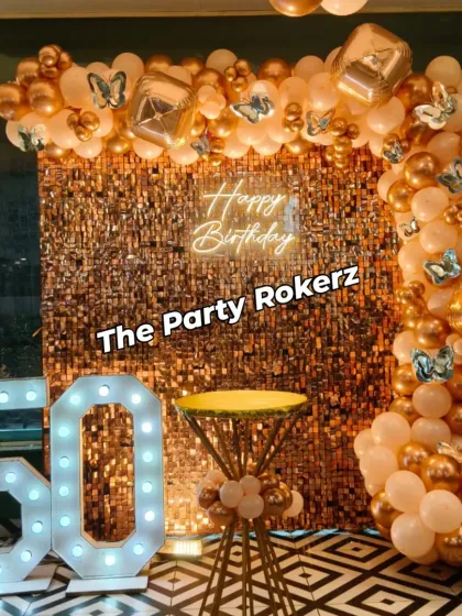 An elegant and celebratory decor for a 50th birthday. This setup features a gold sequin wall, a "Happy Birthday" neon sign, large marquee numbers, and a stylish organic balloon garland in peach and gold tones.
