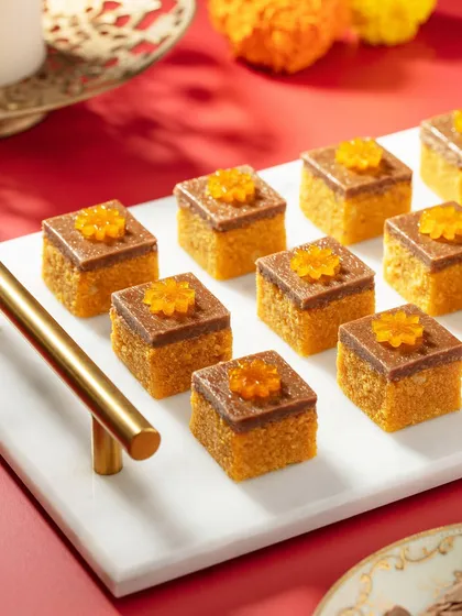A close-up of delicate Motichoor and chocolate layered sweets from the Marriott Diwali collection. The bright red background and clean marble tray make the intricate details and vibrant orange color of the mithai stand out.