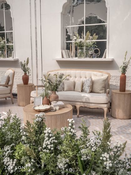 A cozy seating nook with a classic tufted sofa set against a mirrored window, surrounded by lush greenery and delicate floral arrangements.