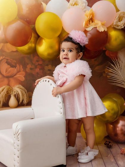 This little girl is ready to celebrate her birthday, standing proudly in her pretty pink dress next to a mini armchair.