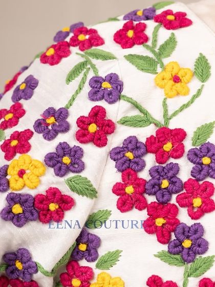 A garden of flowers on a blouse. This white raw silk piece is covered in vibrant, multi-colored floral French knot embroidery, making it a cheerful and eye-catching addition to any festive wardrobe.