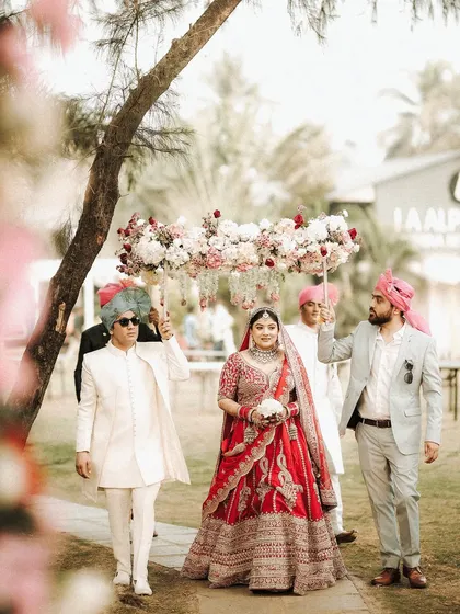 A wide shot of the bridal procession. This image captures the tradition and beauty of the bride's walk to the mandap, surrounded by family in a lovely garden setting.