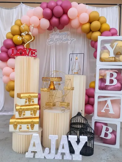A warm-toned birthday balloon ring in pink and gold, arranged with natural wood-finish pedestals and other decorative elements.