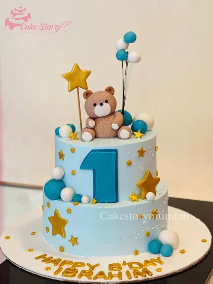 A lovely two-tier cake for a first birthday. The blue cake is decorated with stars, balloons, and a cute fondant teddy bear, creating a dreamy theme for a baby boy.