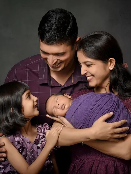 A beautiful family of four admiring their newest member. The older sister gently touches the newborn, wrapped in a vibrant purple swaddle, creating a colorful and heartwarming portrait against a dark canvas.
