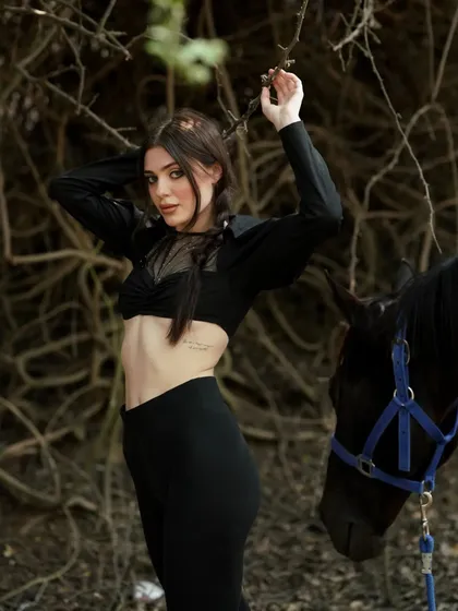 This image captures a moment of connection between the model and the dark horse, set against a natural, textured background.