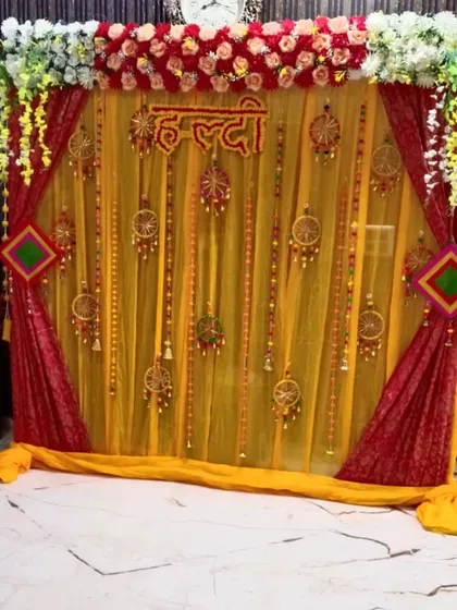 This Haldi backdrop uses a combination of sheer yellow and rich red drapes for a royal look. The decor is completed with floral arrangements at the top, a "Haldi" sign, and decorative hangings for a festive feel.