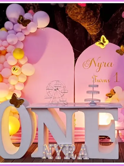 A soft and sweet butterfly-themed first birthday. The pink decor features a large 'ONE' table, butterfly accents, and a 'NYRA' marquee.
