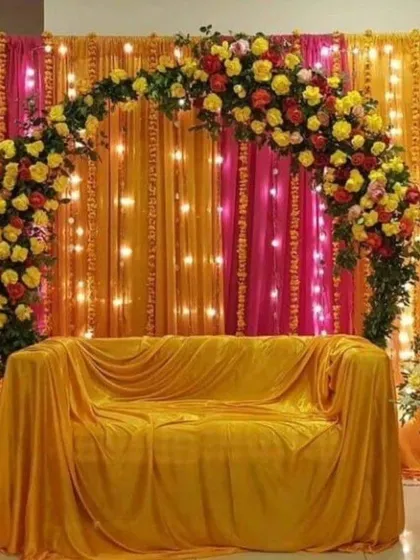 A warm and inviting seating area with a floral arch against a backdrop of orange and pink drapes with fairy lights. The yellow draped sofa is perfect for the bride at her Haldi or Mehandi.