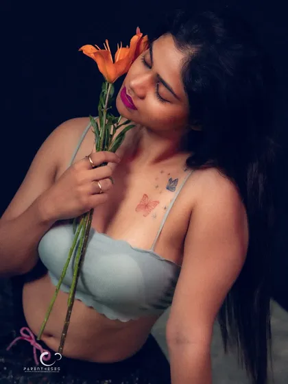 A soft and sensual portrait, with her eyes closed as she holds the flower to her cheek. The butterfly tattoo adds another layer of delicate beauty.