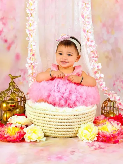 A smiling baby girl in a pink tutu pops out of a basket, surrounded by a beautiful pink floral backdrop.