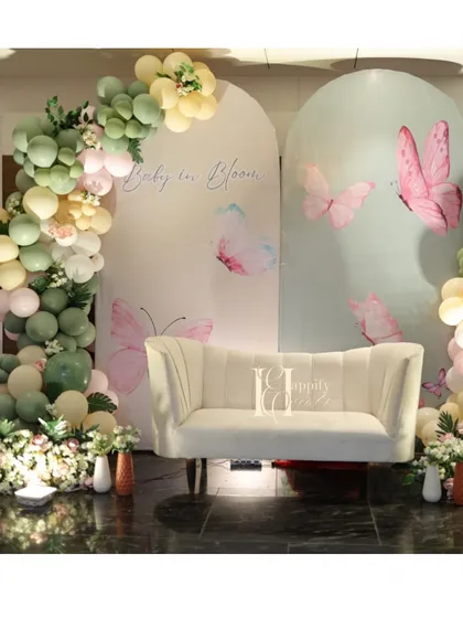 A beautiful "Baby in Bloom" seating area for the parents-to-be. The setup includes two arched backdrops with a soft butterfly print, an organic balloon garland in sage green and pastel pink, and a comfortable white sofa.