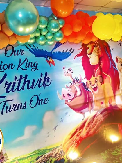 A vibrant Lion King backdrop for a first birthday. The colorful scene from the movie is the centerpiece, framed by a bright orange and yellow balloon garland.