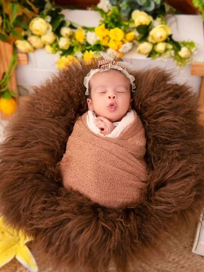 A newborn makes a cute pout while sleeping, wrapped in a brown swaddle and resting in a fur-lined basket. We are always ready to capture these unique and adorable expressions that showcase your baby's budding personality.